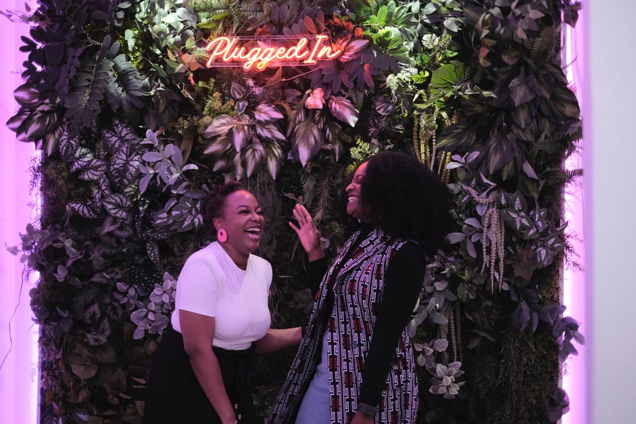 2 women posing in front of a PluggedIn backdrop